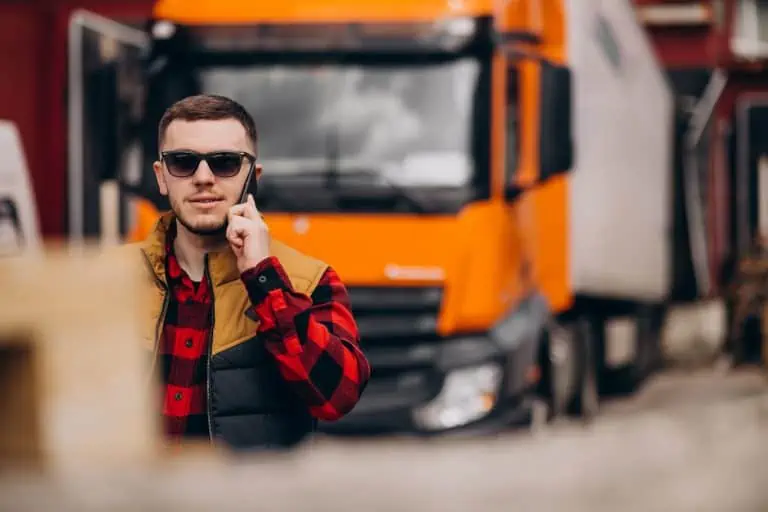 Young male driver standing in front of orange semi truck on the phone.