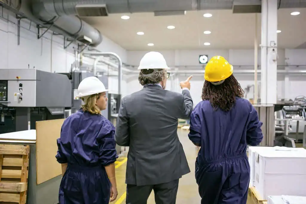 Male engineer and female factory employees in hardhats walking on plant floor and talking, man pointing at equipment and instructing women. Back view. Industrial occupation concept