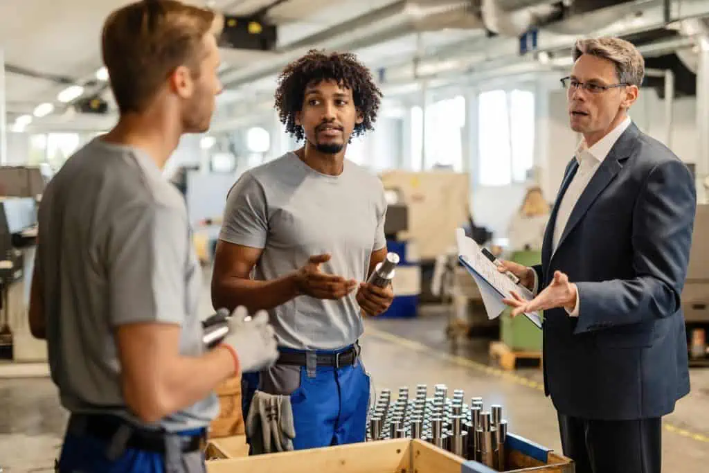 Mid adult manager discussing with metal workers while performing quality control check of manufactured rod cylinders in a factory.
