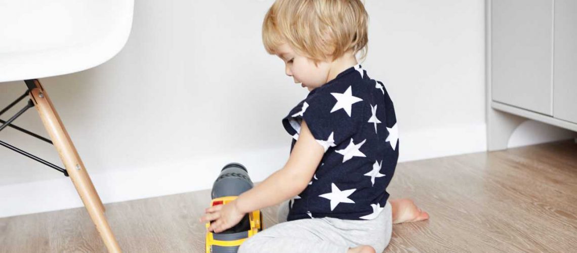 Cute baby boy with blonde hair sitting on wooden floor in his bedroom, holding his favourite toy and smiling. Toddler having fun, playing with yellow plastic truck. Early learning. Developing toys