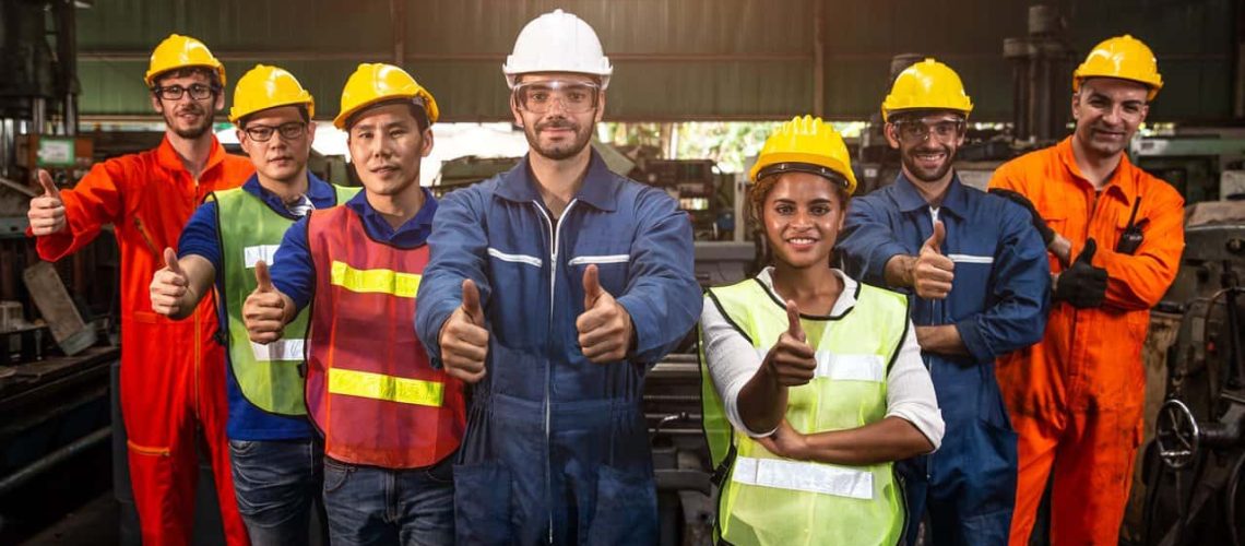 Diverse group in a warehouse in safety equipment giving a thumbs up.