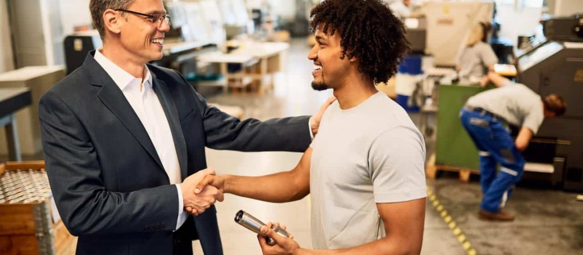 Happy manager shaking hands with African American factory worker in industrial building.