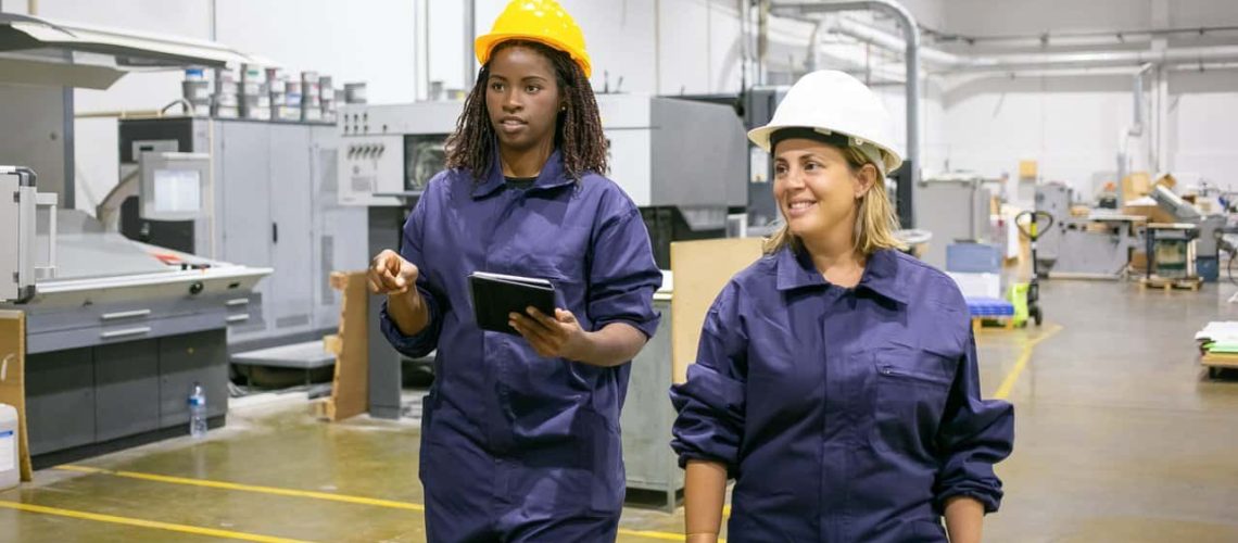 African American worker pointing, talking with colleague and holding tablet. Professional women working at factory and wearing protective uniform. Manufacture, workflow and digital technology concept